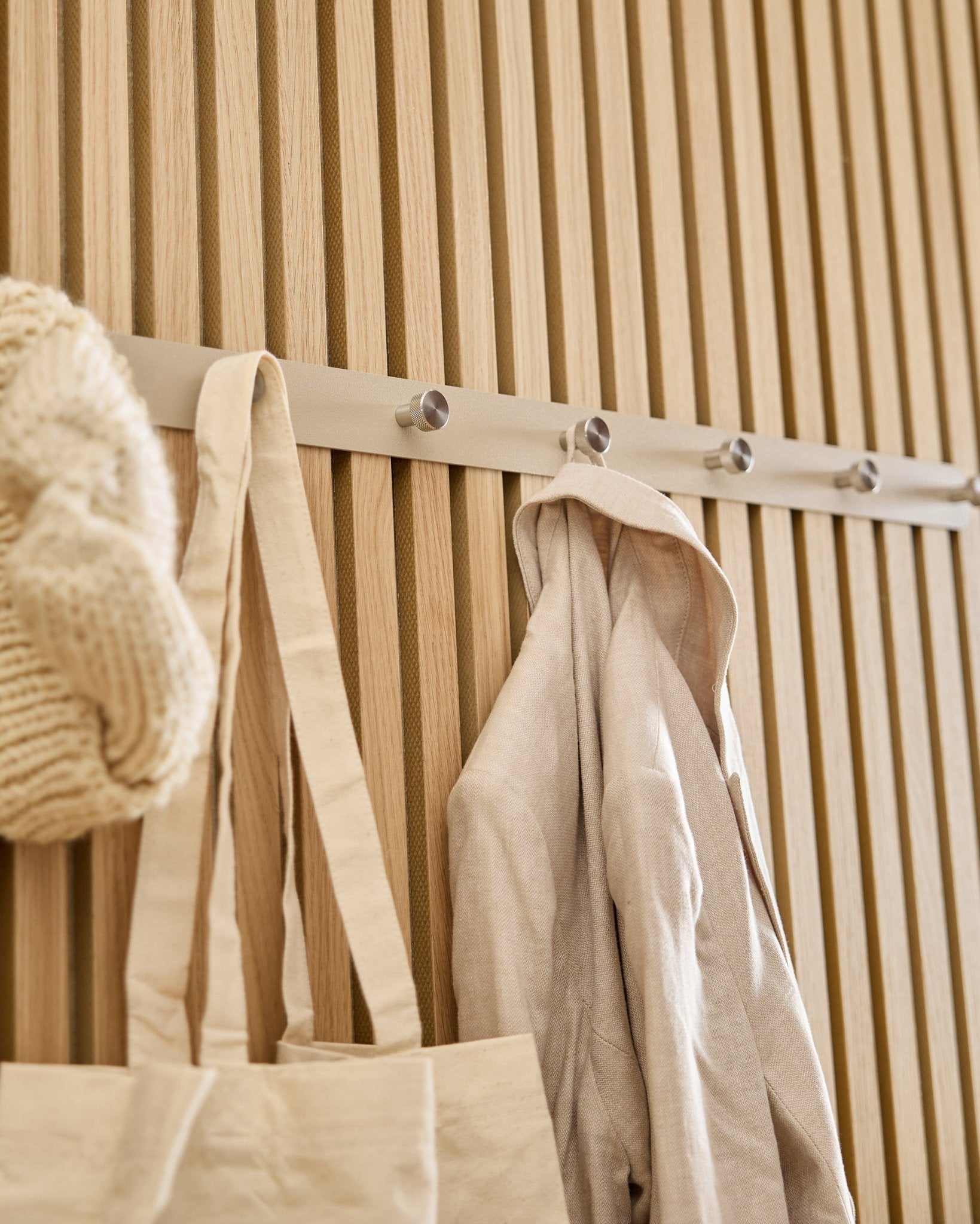 En beige totebag, strikhue og hørskjorte hænger på WoodUpp Norr knagerække (Beige/rustfrit Steel, X7), monteret på et lyst Akupanel lamelpanel i træ.